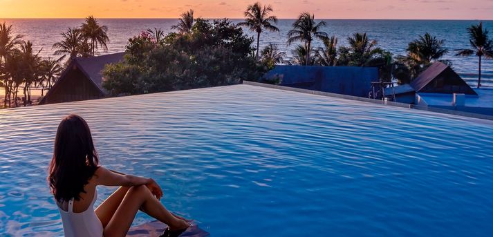 Frau sitzt an einem Swimmingpool und blickt auf das Meer und die untergehende Sonne