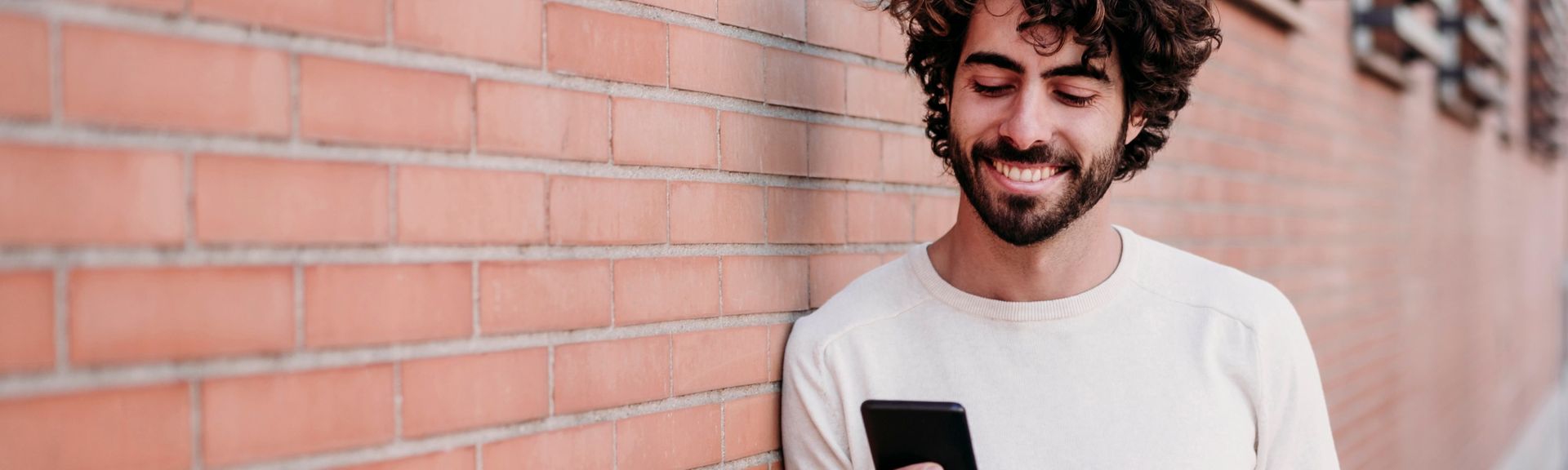 Junger Mann lehnt lässig an einer Hauswand und blickt lächelnd auf sein Mobiltelefon