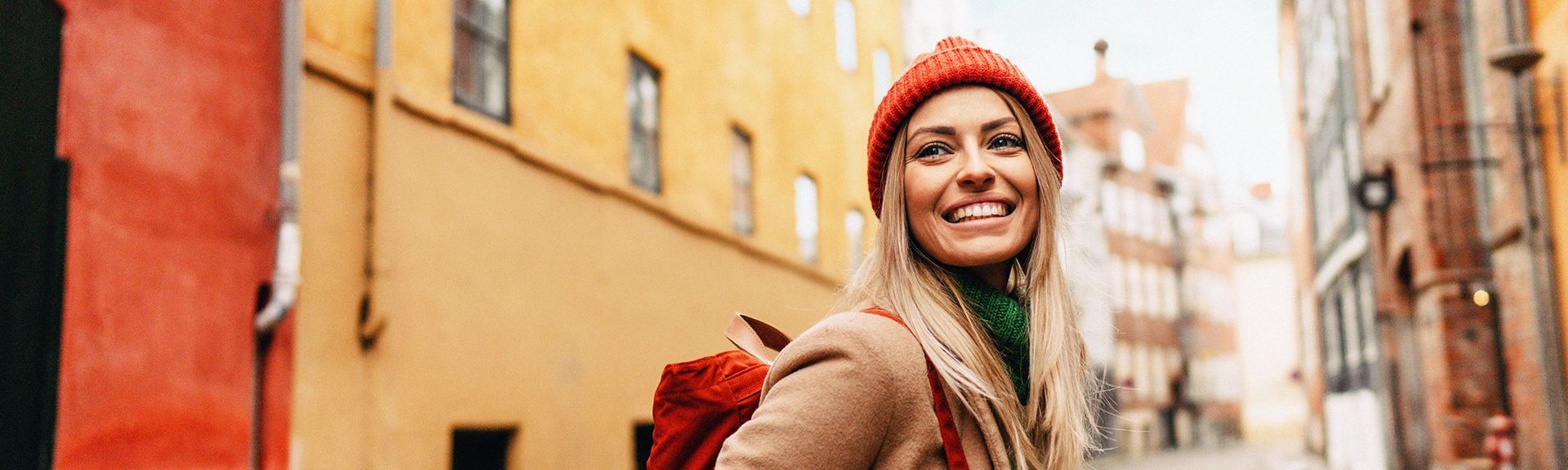 Frau mit Rucksack und Mütze steht in einer kleinen Gasse und lächelt