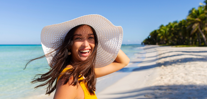 Eine Frau mit Hut läuft lächelnd an einem sonnigen Palmenstrand entlang