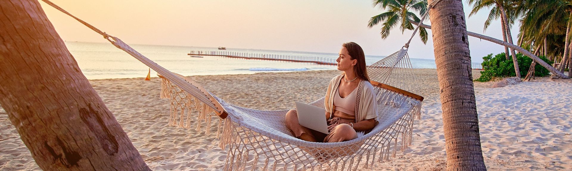Frau mit Laptop sitzt in einer Hängematte am Strand und blickt aufs Meer