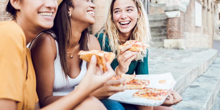 Drei Freundinnen sitzen in einer italienischen Stadt und essen lachend eine Pizza