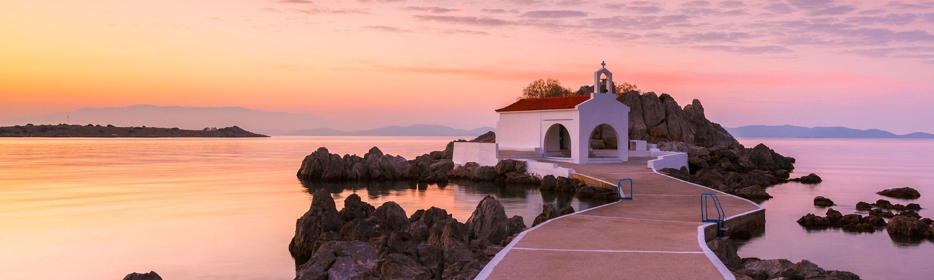 Blick auf die Chios Kirche. Im Vordergrund ist ein Steg der zur Kirche führt und im Hintergrund sieht man Meer und Himmel im Sonnenuntergang.